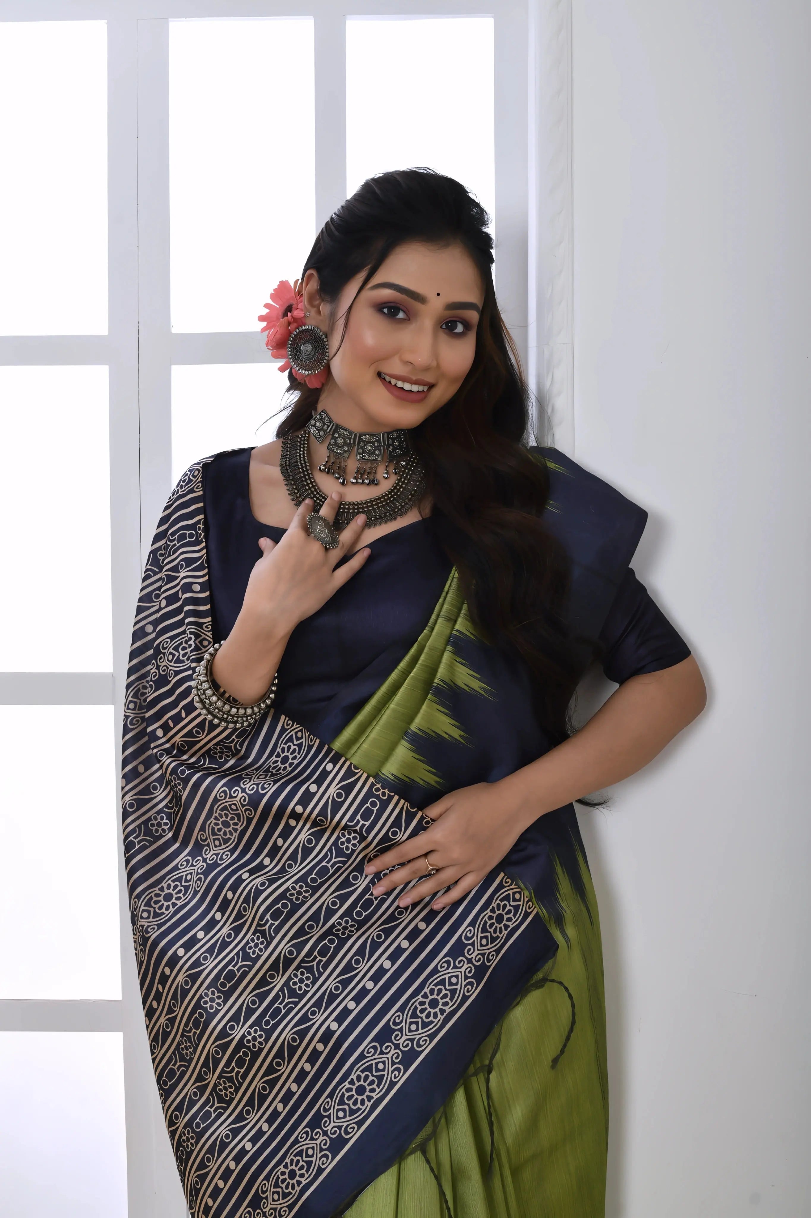 A woman models a striking Pista Green Soft Tussar Silk Saree. The saree features a bold black/navy zigzag or V-shaped pattern along the border and transitions on the body. The pallu has a wide panel of dense tribal or Ikat-style geometric print in light grey and white on a dark background. She wears a black/navy blouse and chunky oxidized silver jewelry, posing indoors by a large arched window.